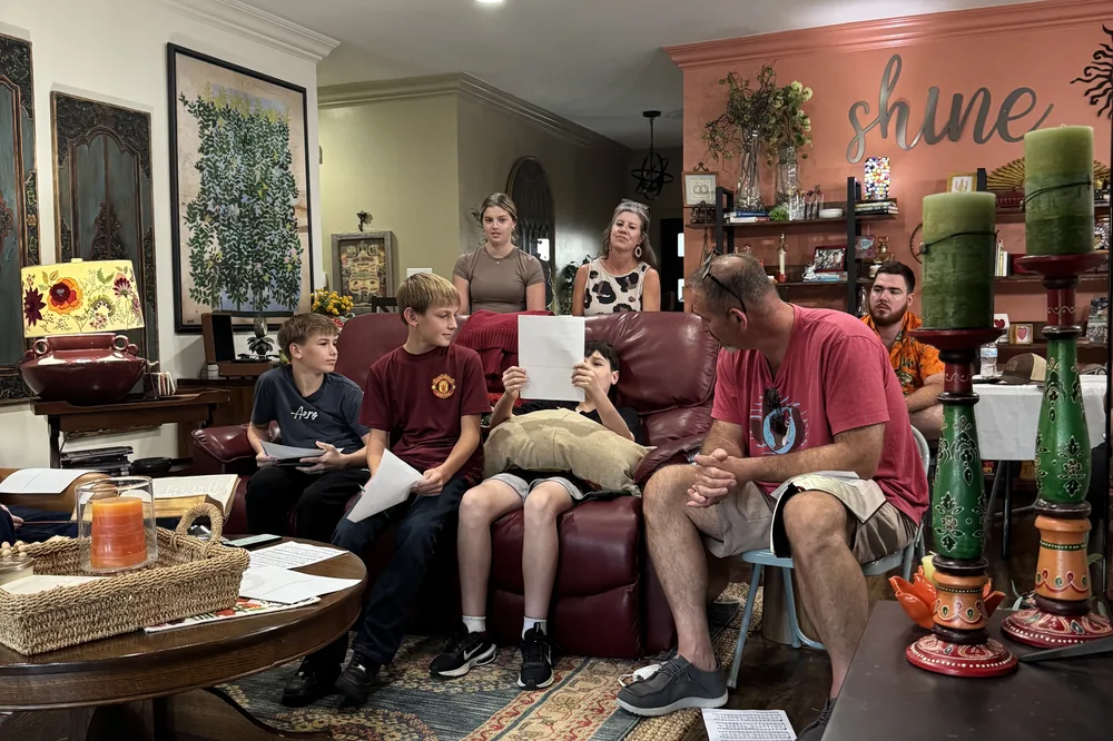 Students and adults gathered in a living room, song sheets spread on the table
